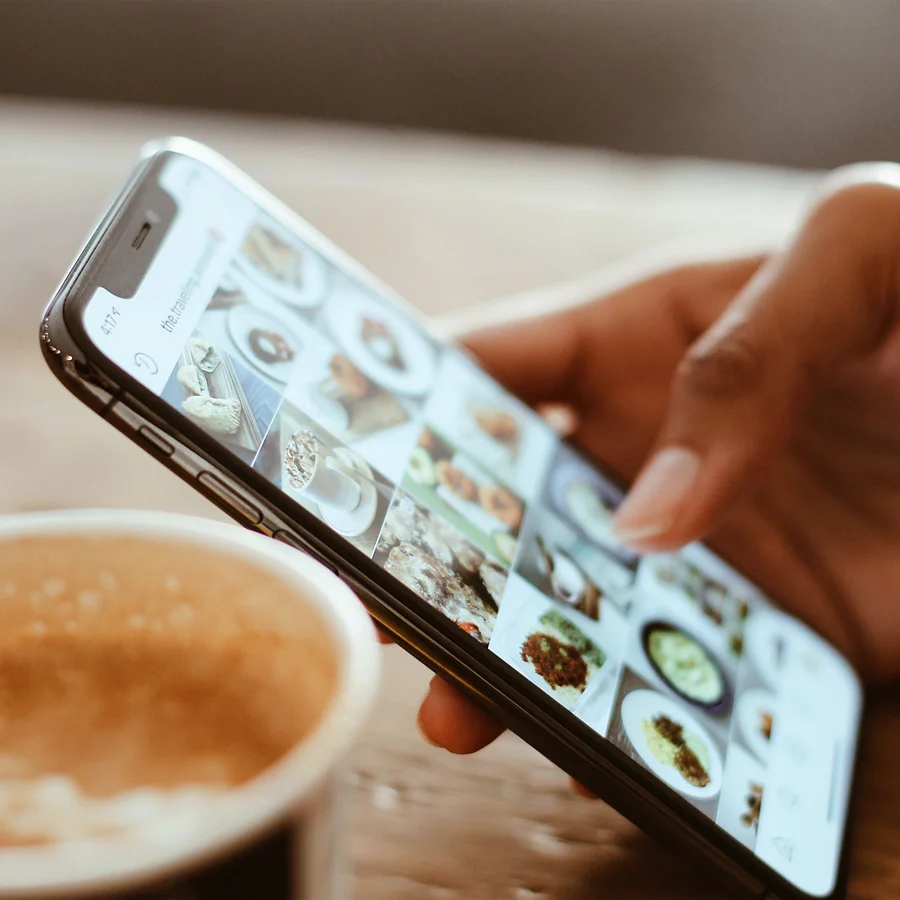 Eine Hand mit einem Smartphone, auf dem ein Instagram-Feed zum Thema Essen angezeigt ist und ein Kaffee links davon